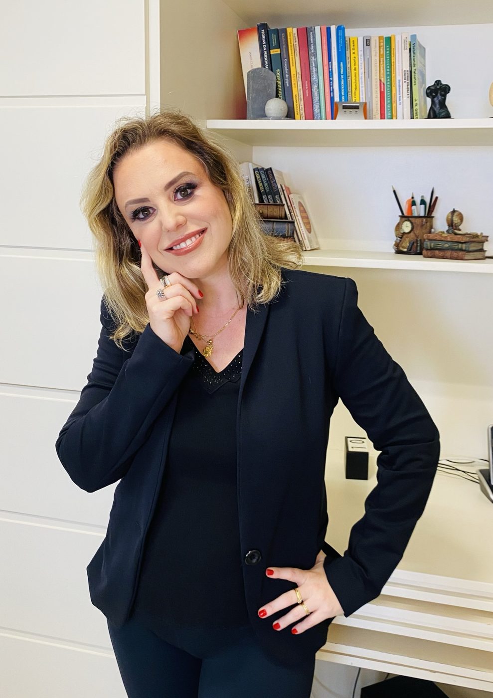 Dra. Vanessa Heck de blazer preto, com a mão no rosto, diante da estante do consultório